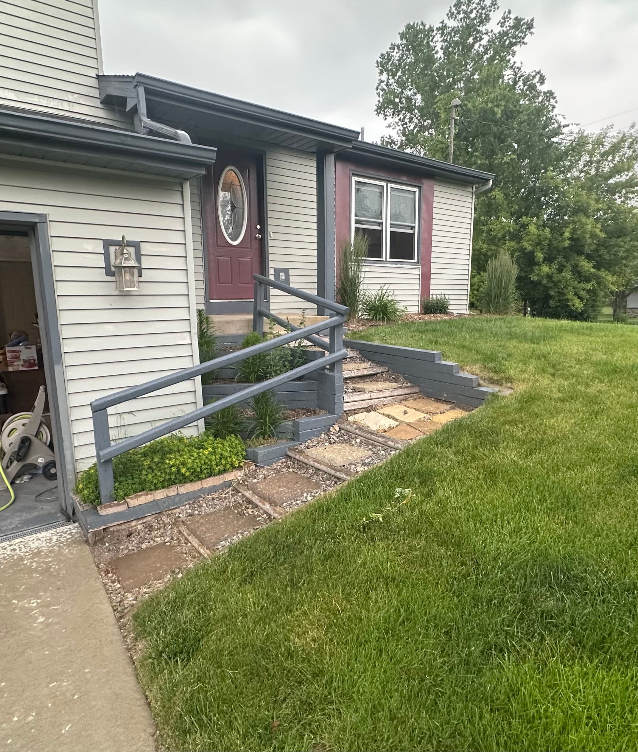 Front entrance of a house with stairs, decorative plants, and a gray railing on a grassy lawn.