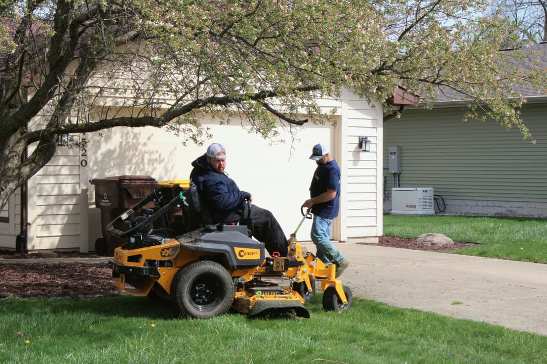 Two landscapers using a riding lawn mower to maintain a residential yard in spring.