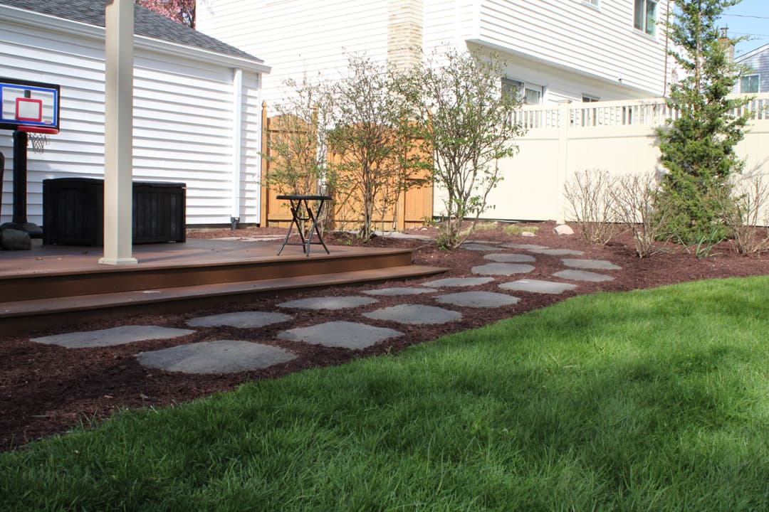Backyard landscape with stone pathway, lawn, and wooden deck in suburban setting.