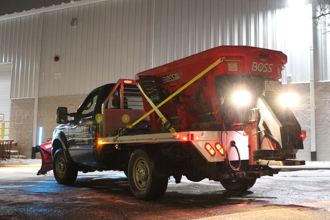 Snow plow truck with a red BOSS spreader, illuminated at night on a snowy street.