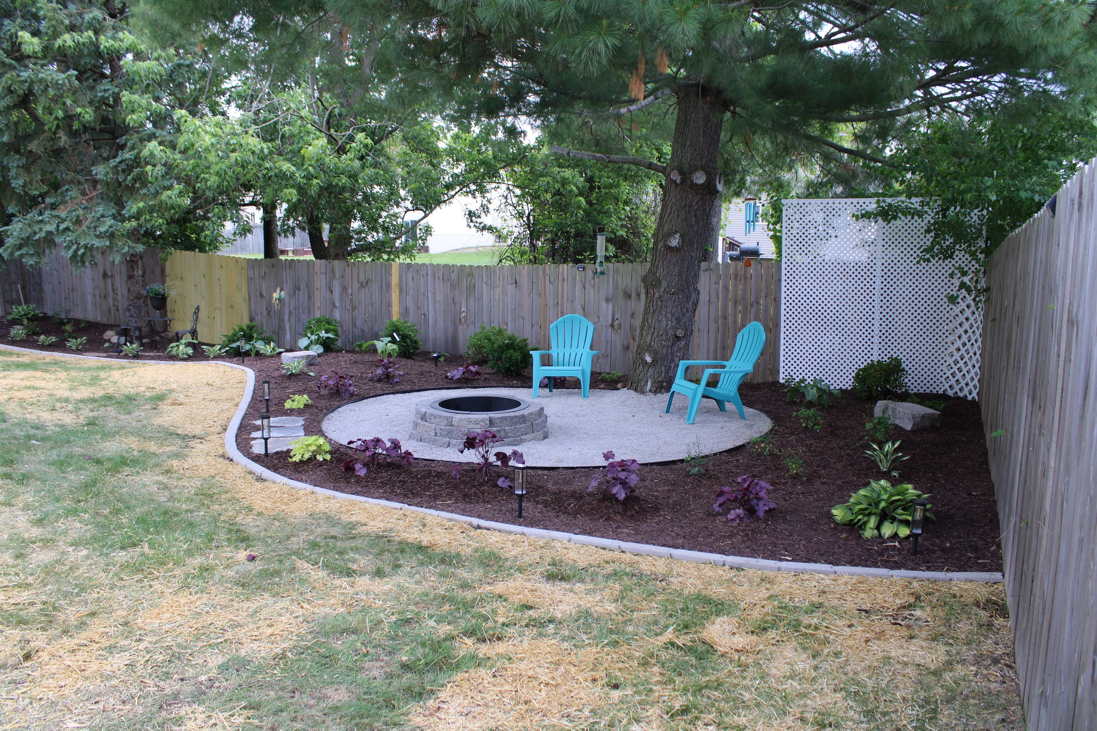 Cozy backyard fire pit area with blue chairs, surrounded by mulch and greenery.