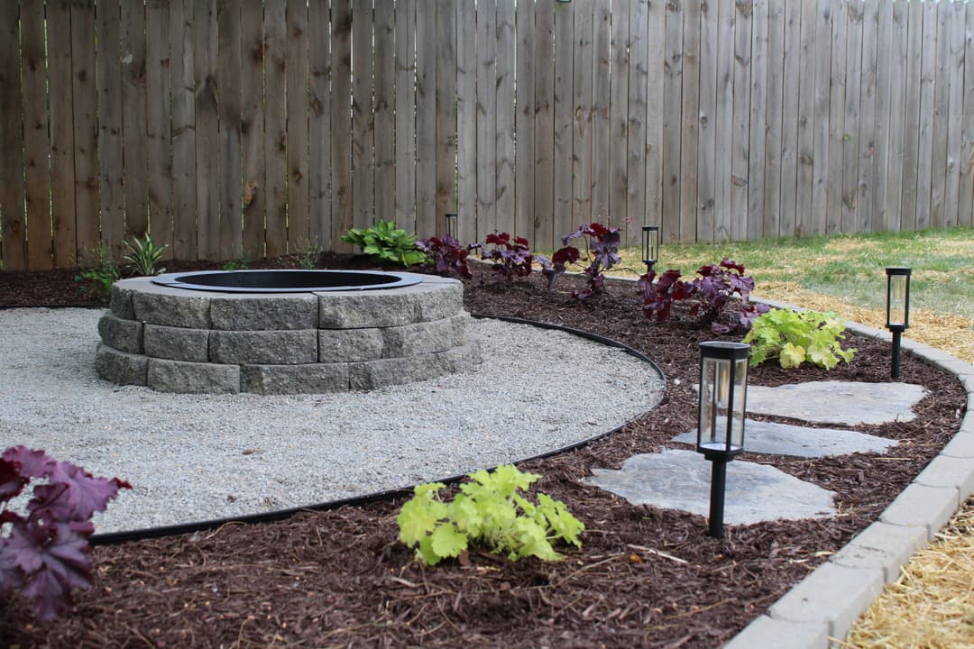 Stone fire pit surrounded by gravel, plants, and solar lanterns in a landscaped backyard.