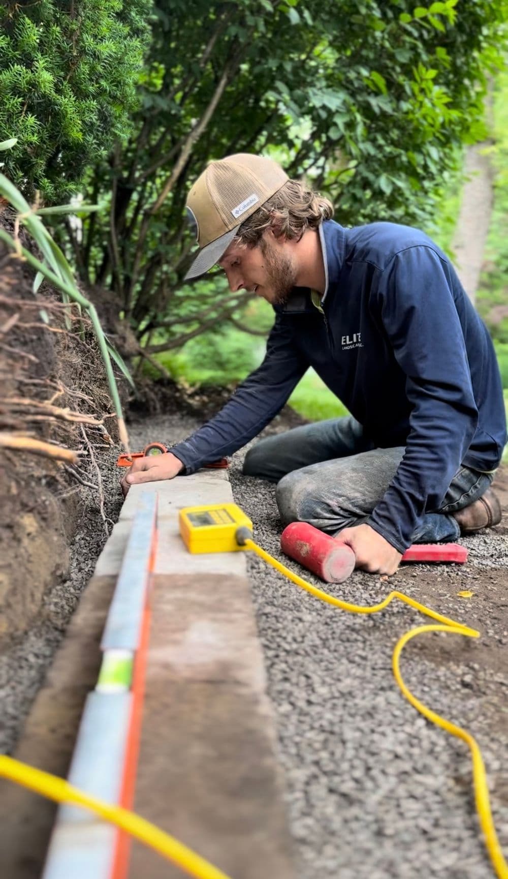 Landscape worker leveling ground with a laser tool in garden setting, surrounded by greenery.