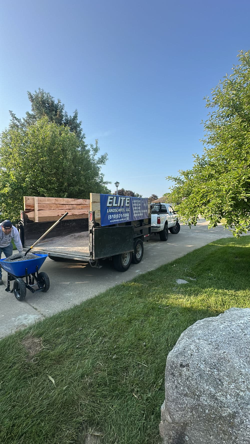 Landscaping crew working near truck with tools on residential driveway, green trees in background.