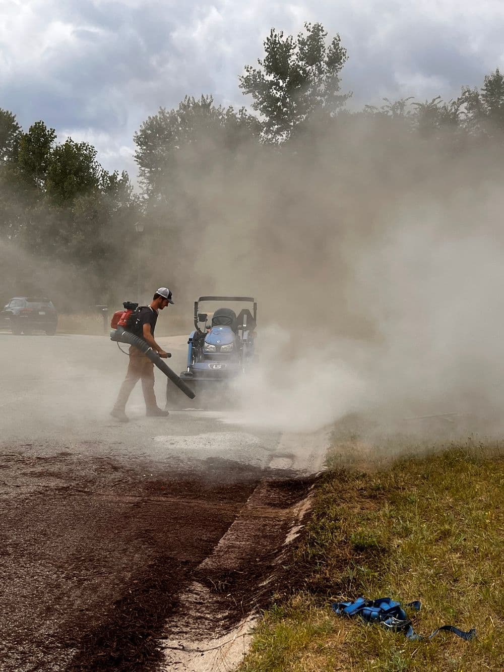 Worker using a leaf blower on a smoky road with machinery in the background.