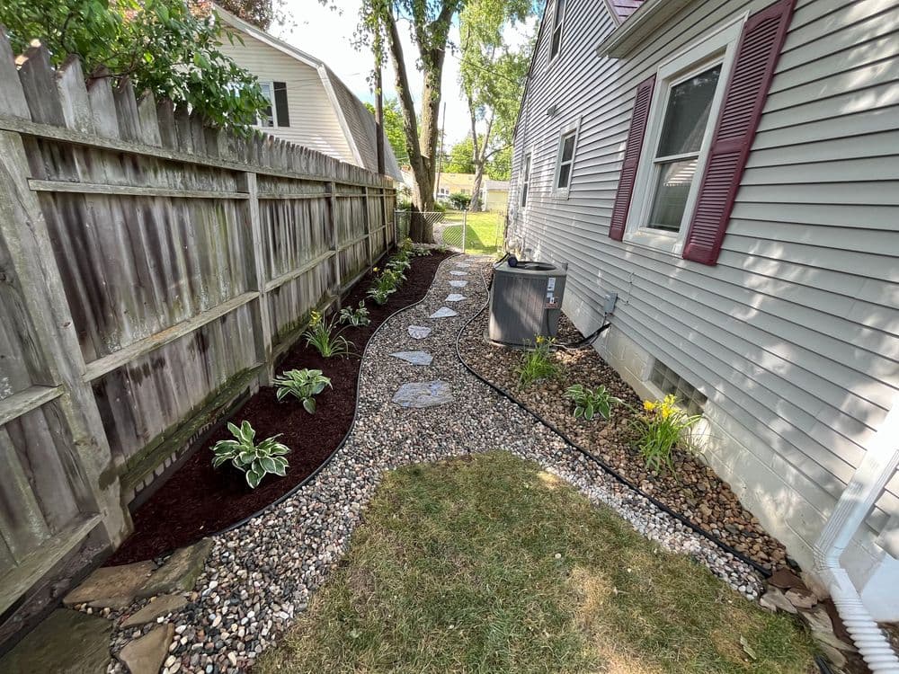 Landscaped side yard with stone path, flower beds, and air conditioning unit beside house.