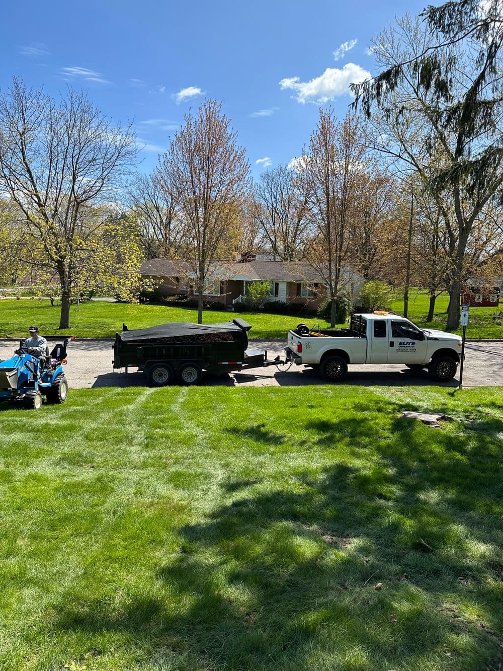 White truck with trailer parked on green lawn under blue sky and trees, residential home in background.