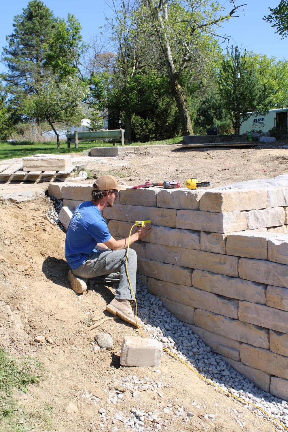 Man measuring a stone wall during landscaping project with tools and greenery in background.