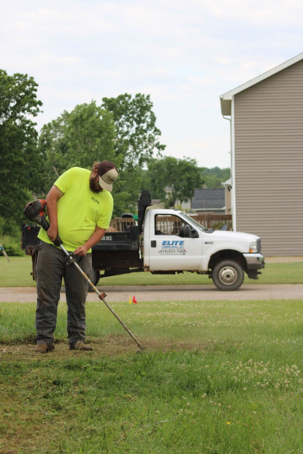 Landscaper using a string trimmer on grass, with a service truck parked nearby.