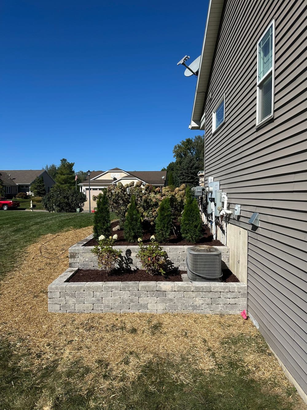 Landscaped garden bed with shrubs and air conditioning unit beside a home on a sunny day.