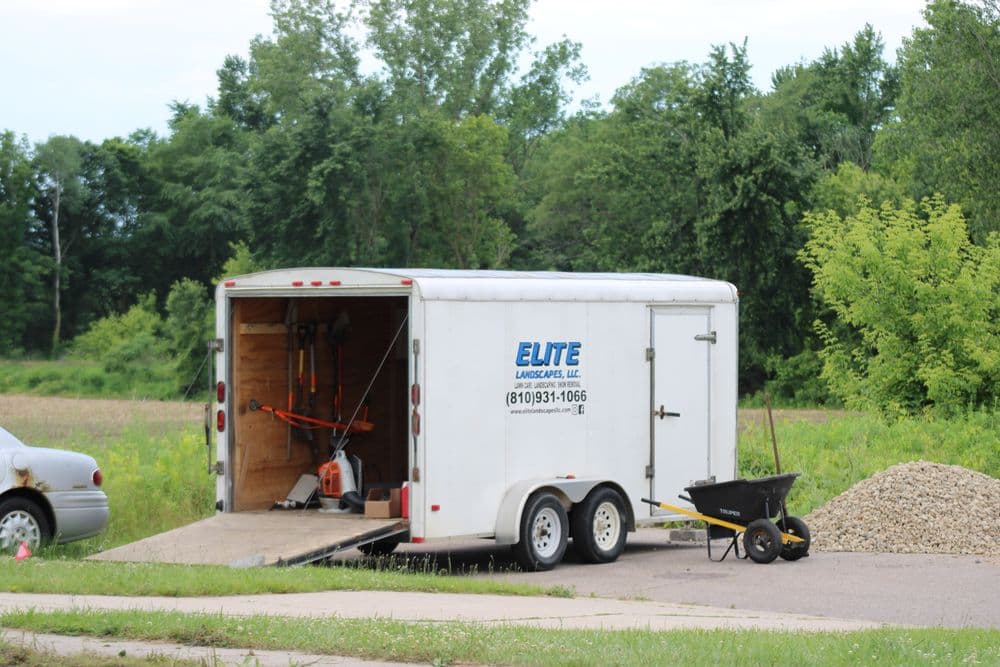 White trailer with landscaping equipment parked beside a gravel area and a wheelbarrow nearby.