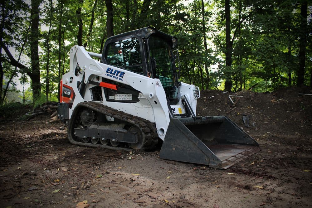 White compact track loader on construction site surrounded by trees and dirt.