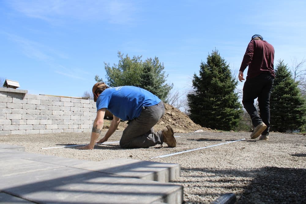 Workers preparing a gravel base for landscaping in a sunny outdoor setting with trees.