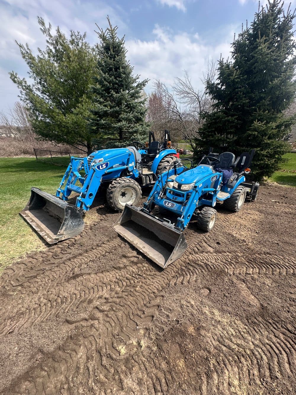 Two blue LS tractors with front loaders parked on freshly tilled soil surrounded by trees.