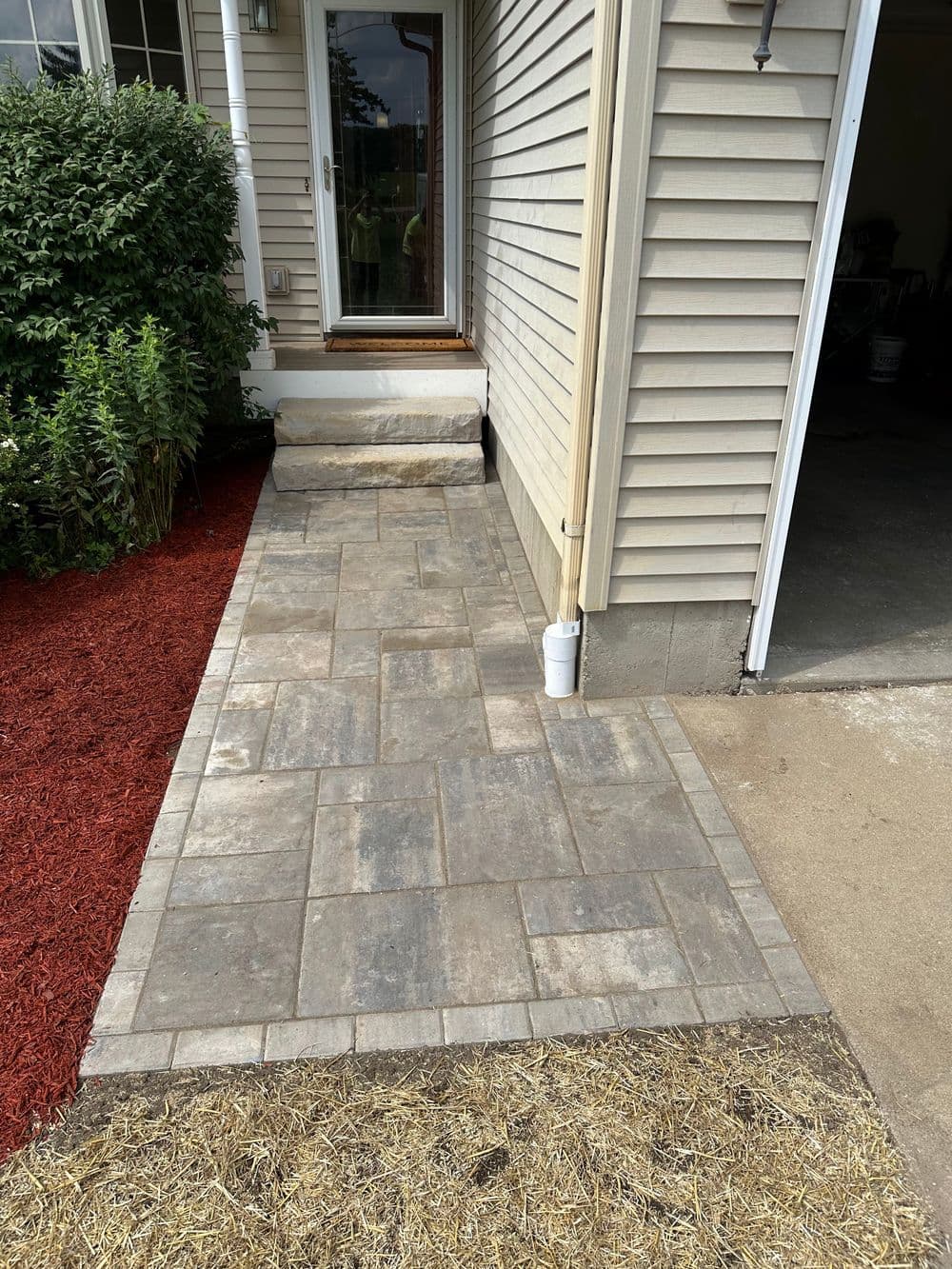 Newly paved stone walkway from house entrance to garage, bordered by landscaping and red mulch.