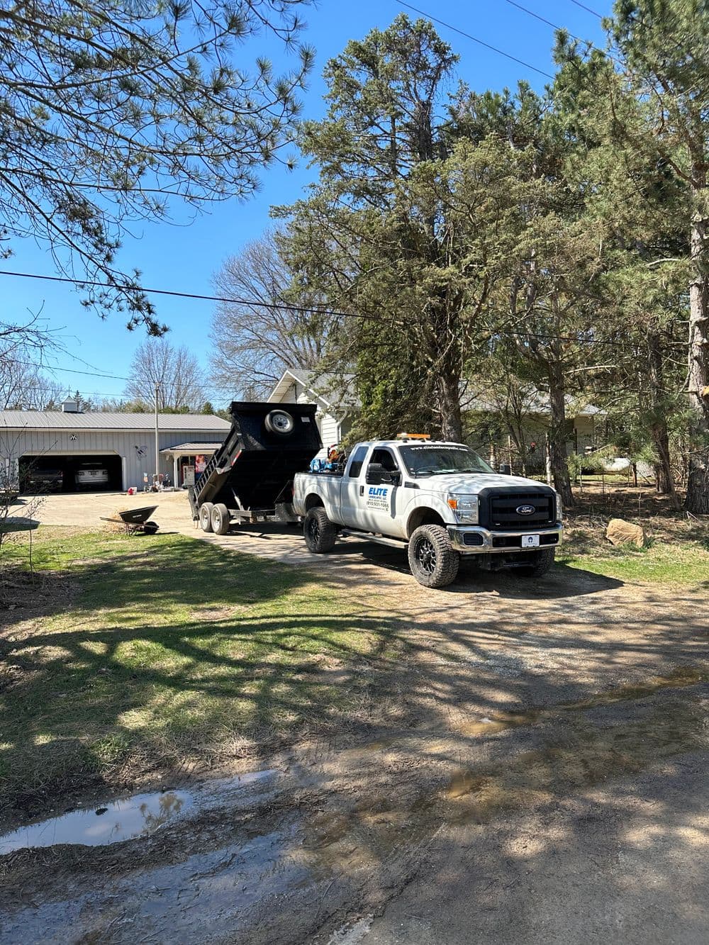 Dump truck on a residential road with trees and a house in the background, under a clear blue sky.