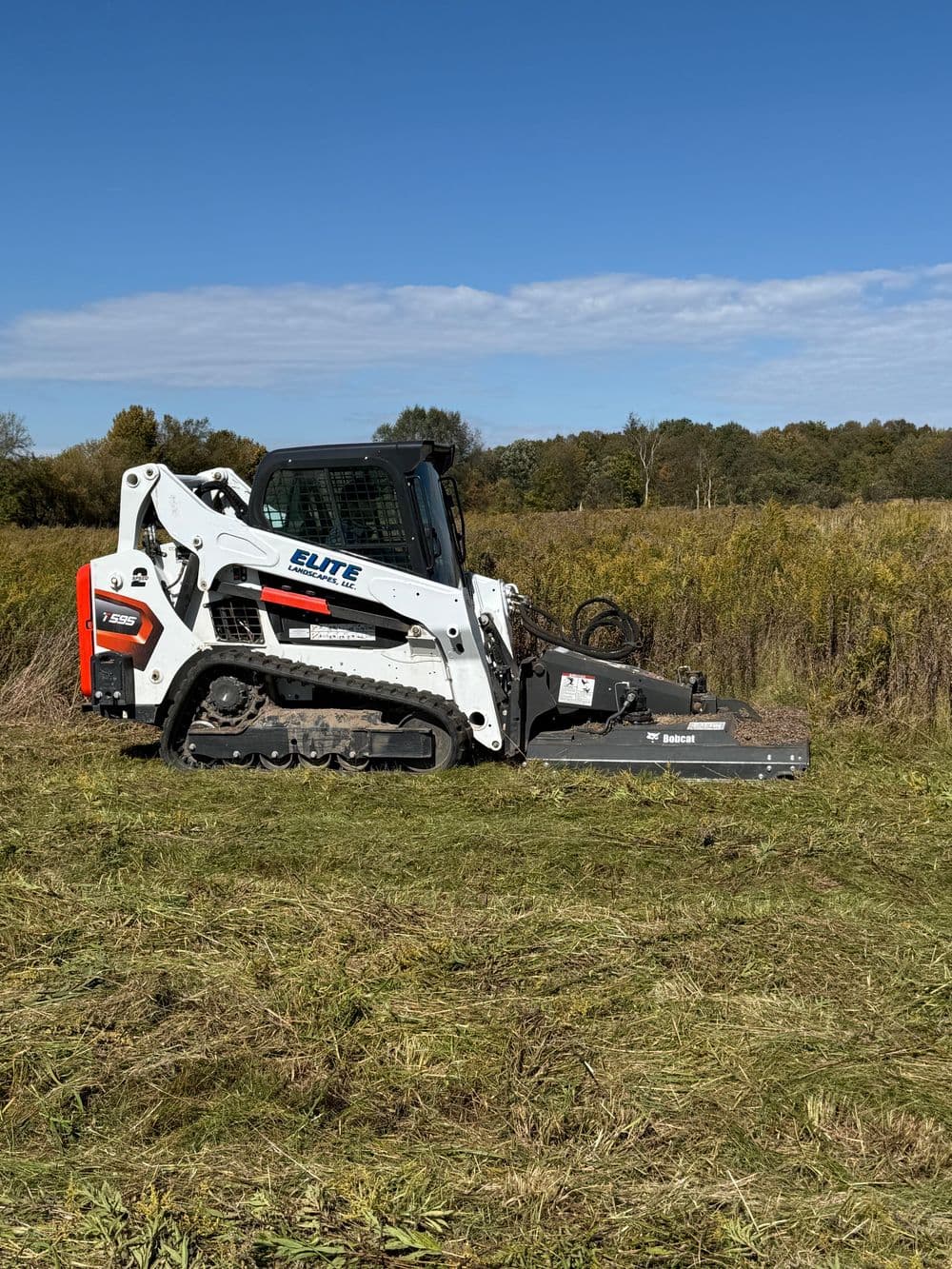 Elite skid steer loader with mower attachment clearing grass in a field under a blue sky.