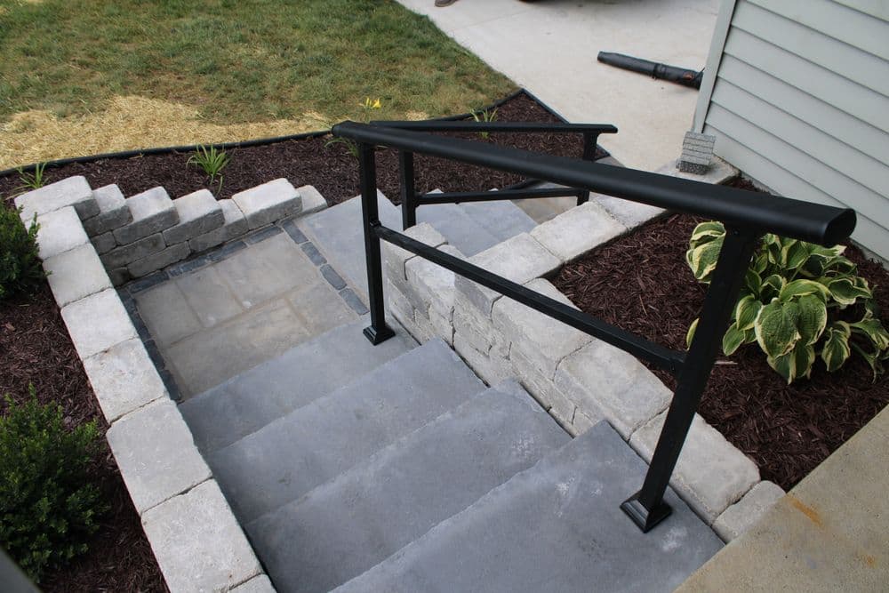 Concrete steps with black handrail leading to a home entry, surrounded by landscaped garden.