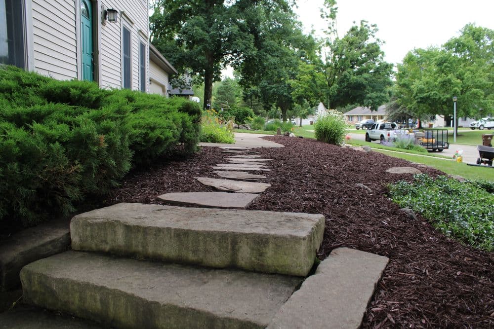 Stone pathway leading to a house, surrounded by mulch and lush greenery in a residential area.