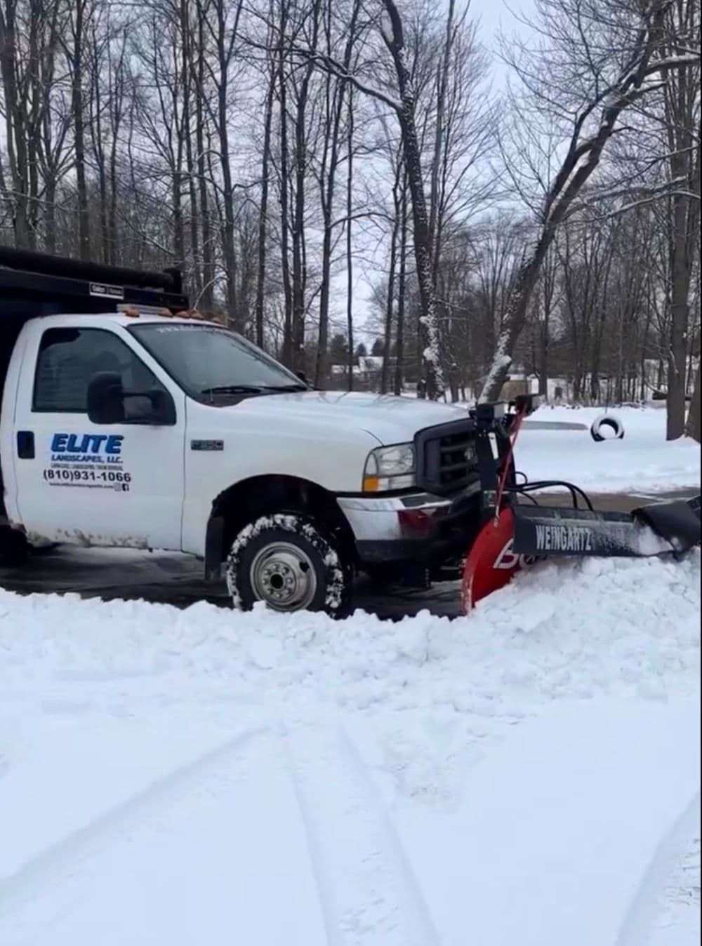 White snow plow truck clearing snow in a winter landscape with bare trees.