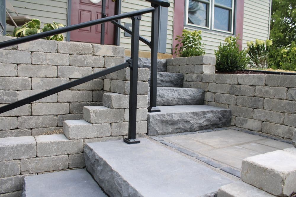 Stone steps with a black railing leading to a home's entrance, featuring landscaping.