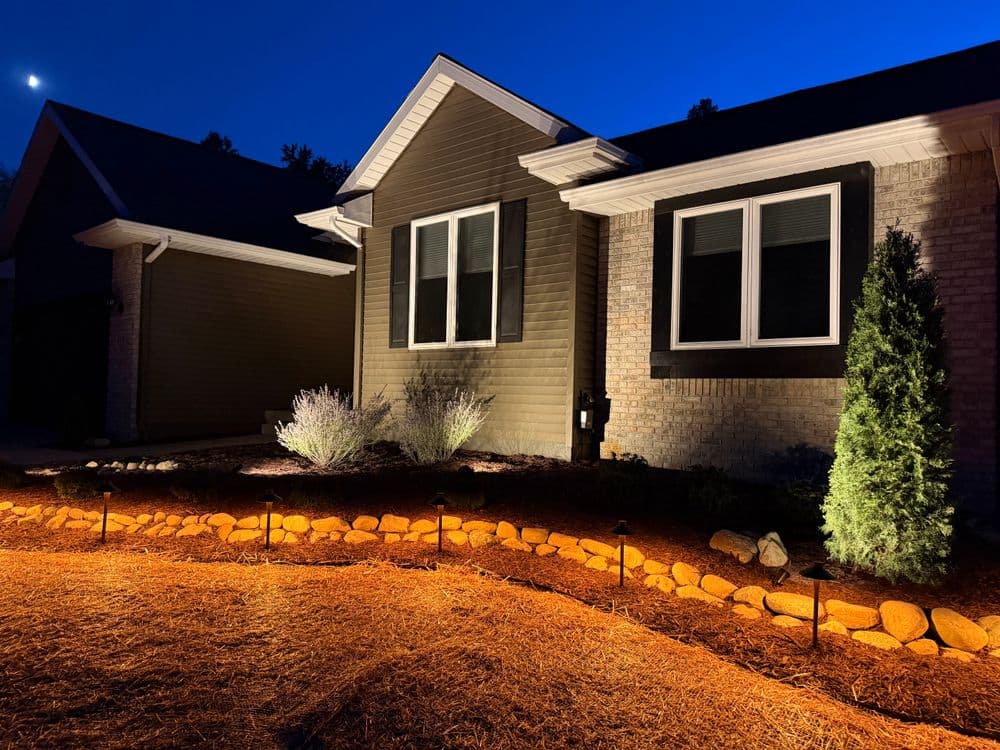 Illuminated modern home exterior with landscaped rocks and shrubs at night.