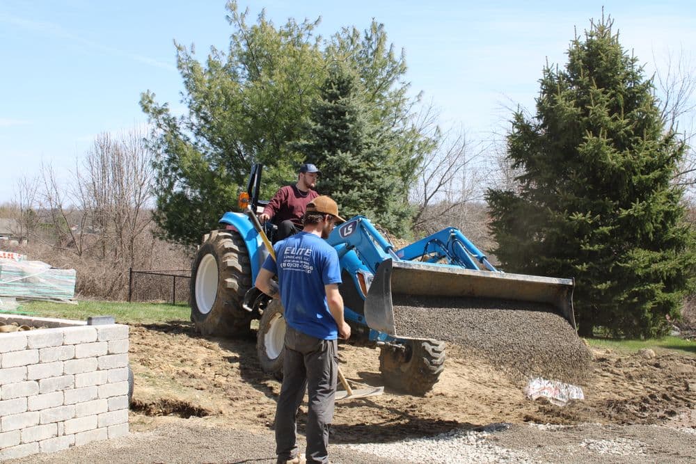 Man operating a blue tractor while another person supervises gravel placement in a yard.