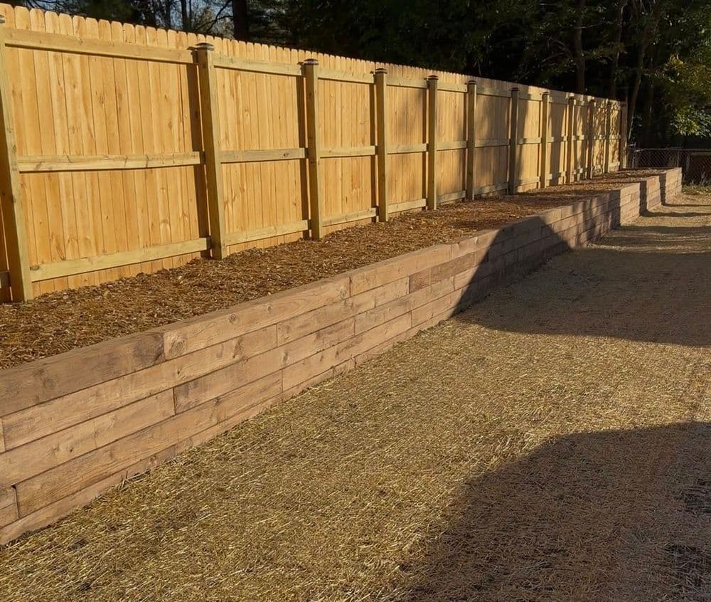 Wooden fence with raised garden beds and mulch in a sunny backyard setting.