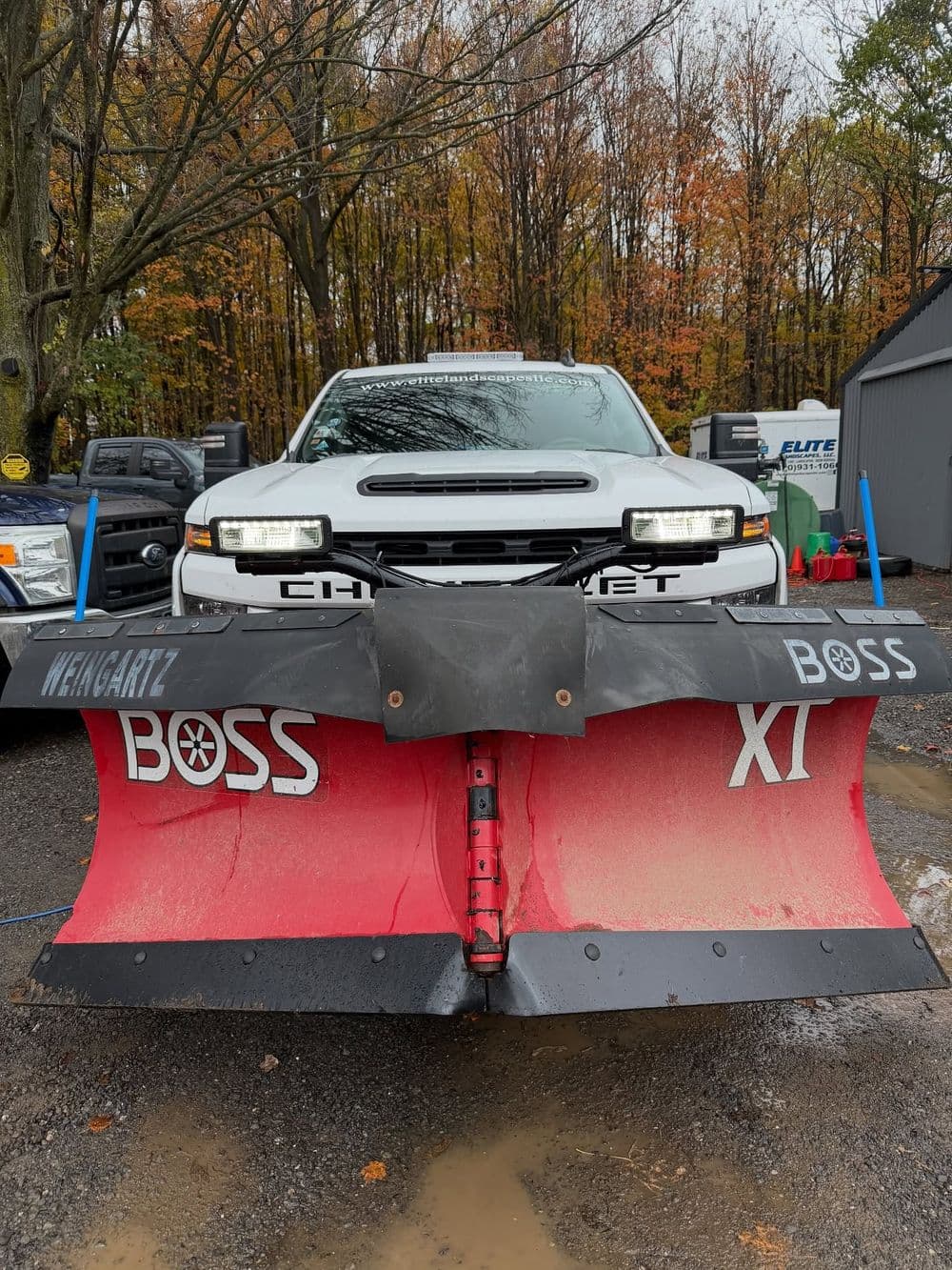White truck with a red snow plow attachment in a forested area during fall.