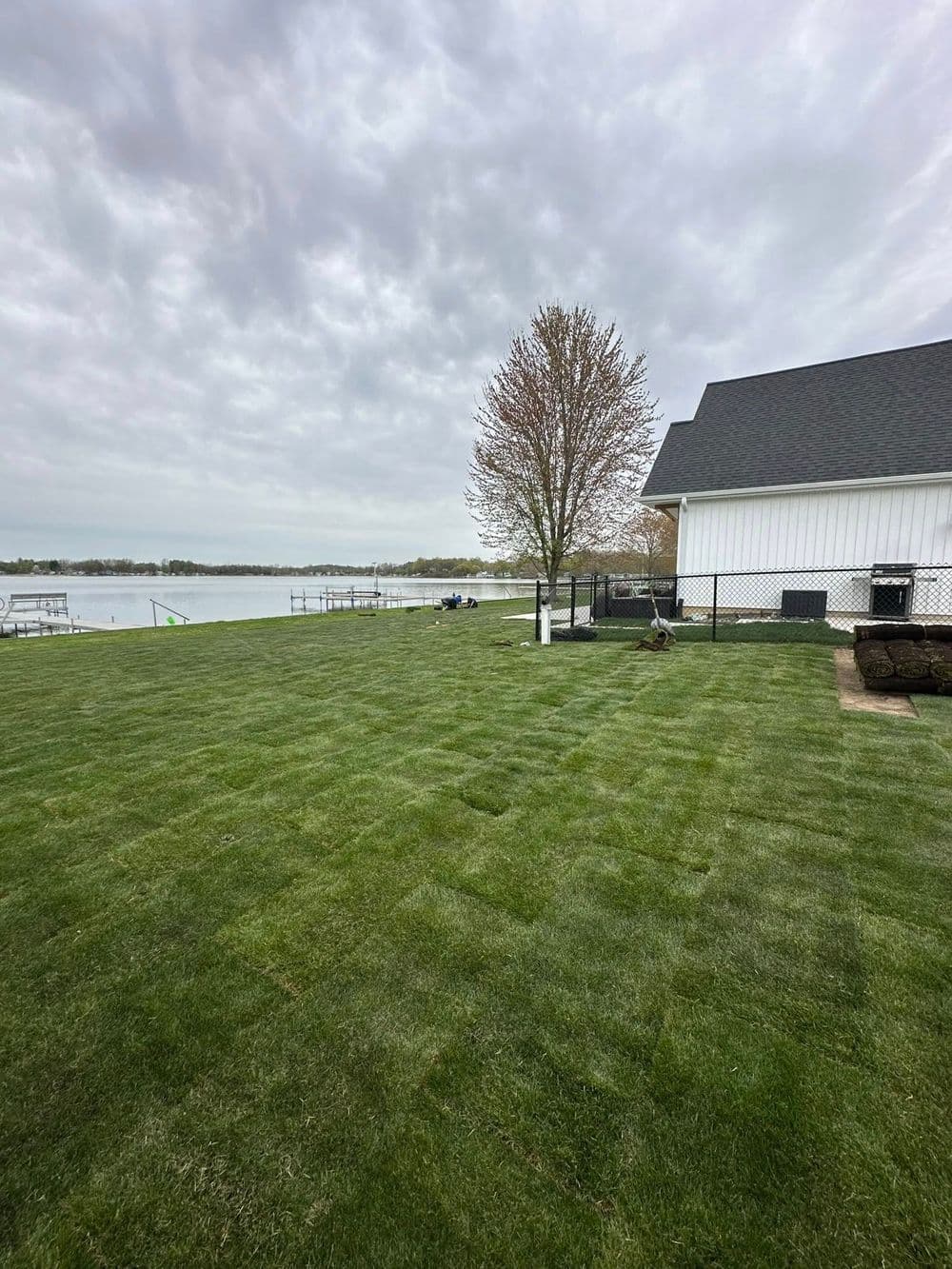 Lush green lawn by a lake with a house and tree under a cloudy sky.