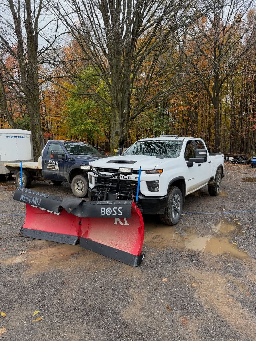 White pickup truck with a red and black snow plow attachment in a forested area.
