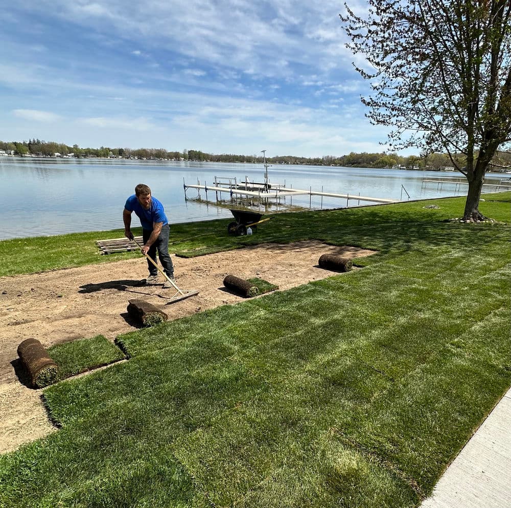Person installing sod in a sunlit backyard with a white fence. Green grass rolls visible.
