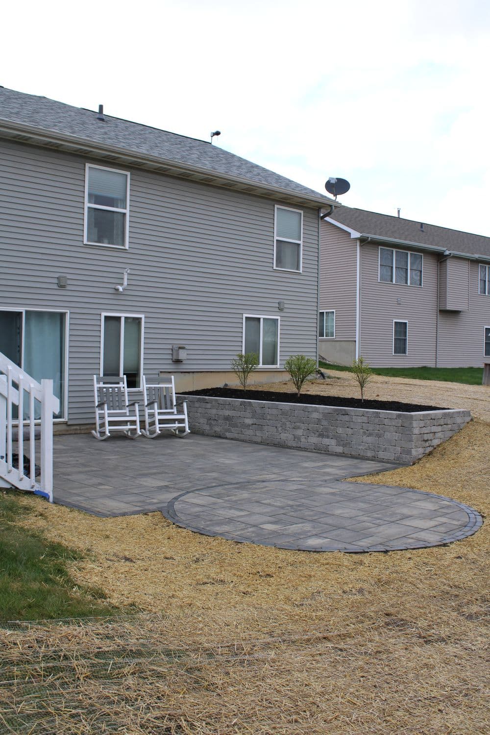 Patio area with stone wall and rocking chairs beside a grassy yard and apartment building backdrop.