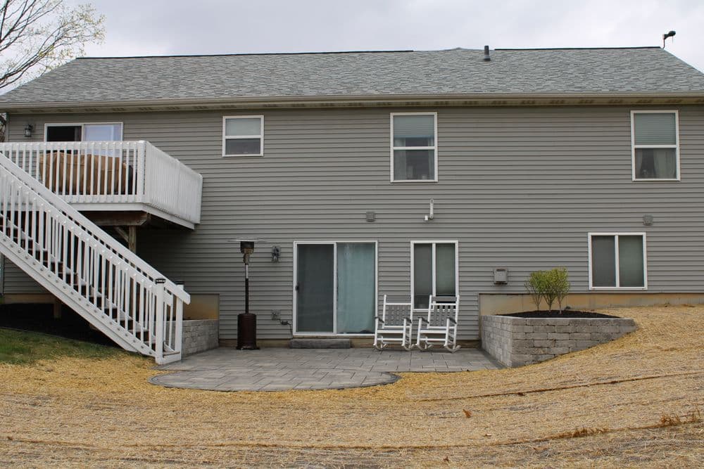 Modern gray house exterior with patio, rocking chairs, and white stairs. Garden area visible.