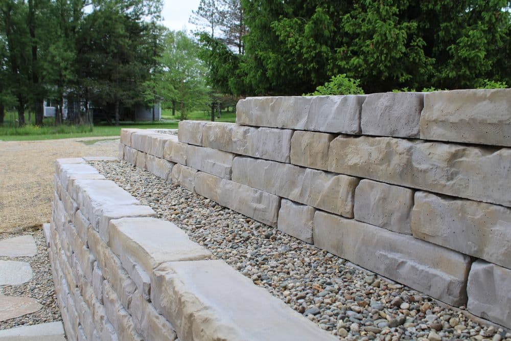 Retaining wall made of layered stone with gravel landscaping and greenery in the background.