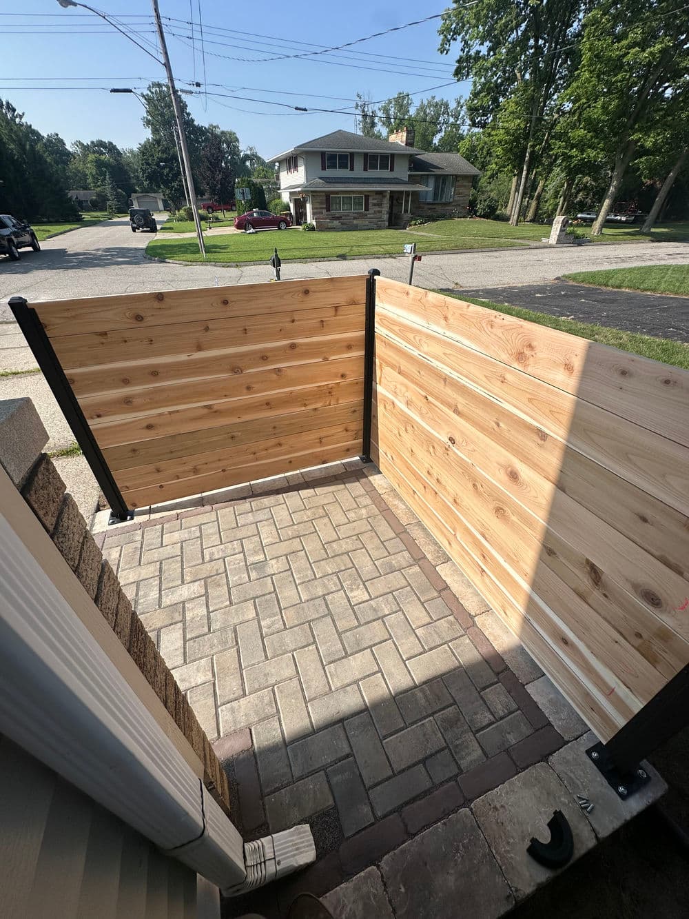 Wooden privacy fence enclosure with brick pavers, residential neighborhood view in background.