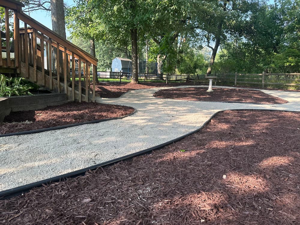 Landscape design featuring a winding gravel path with red mulch and wooden deck under trees.