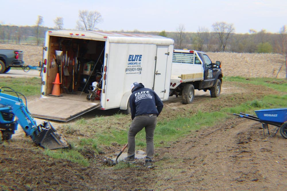 Worker digging near landscaping truck and trailer on a rural site with tools visible.