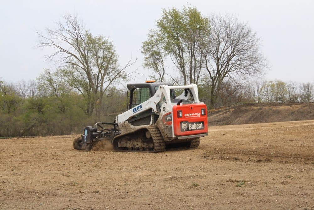 Bobcat skid steer loader landscaping a dirt field with trees in the background.