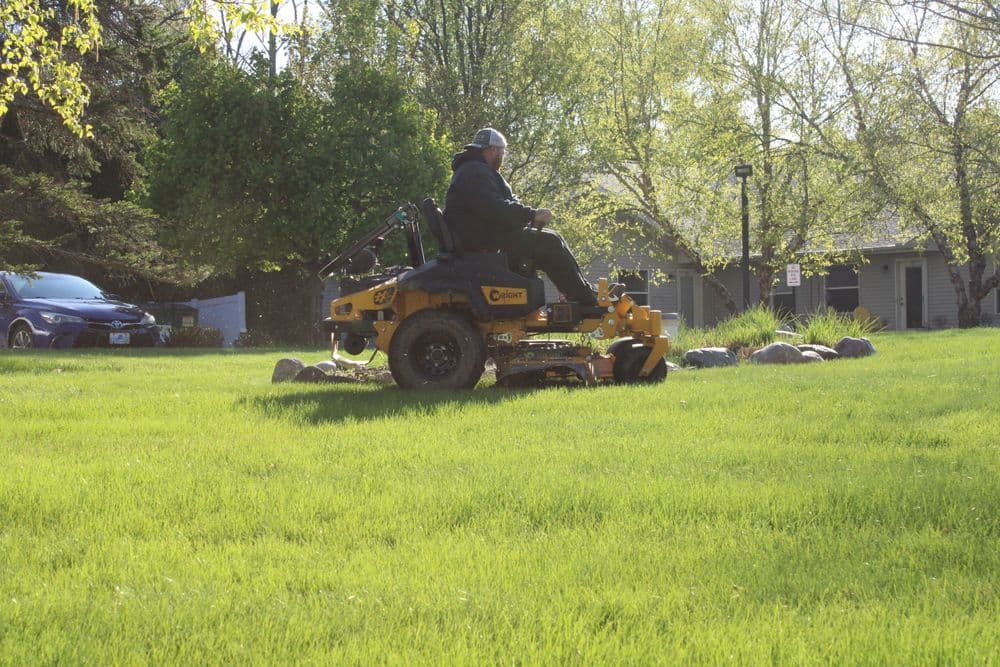 Person mowing a lush green lawn with a riding lawn mower on a sunny day.